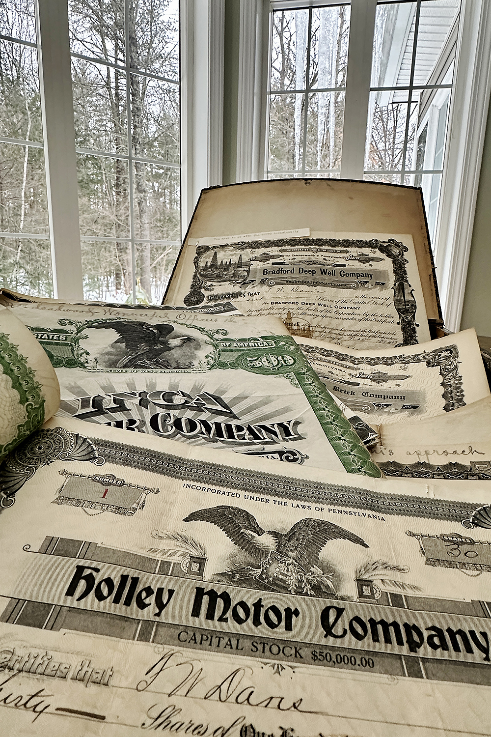 A wooden box opened to vintage stock certificates including Holley Motor Company and Bradford Deep Well Company, near a window in winter light
