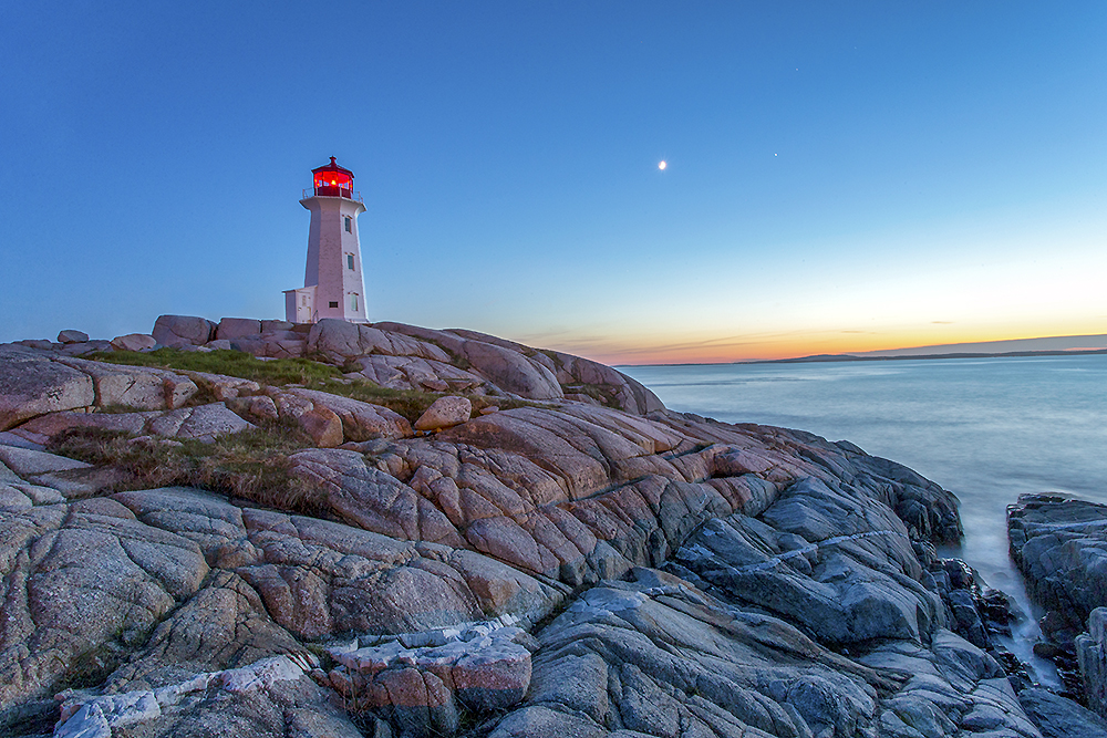 Peggy’s Cove lighthouse illuminated at dusk above calm Atlantic water and smooth granite under a soft twilight sky