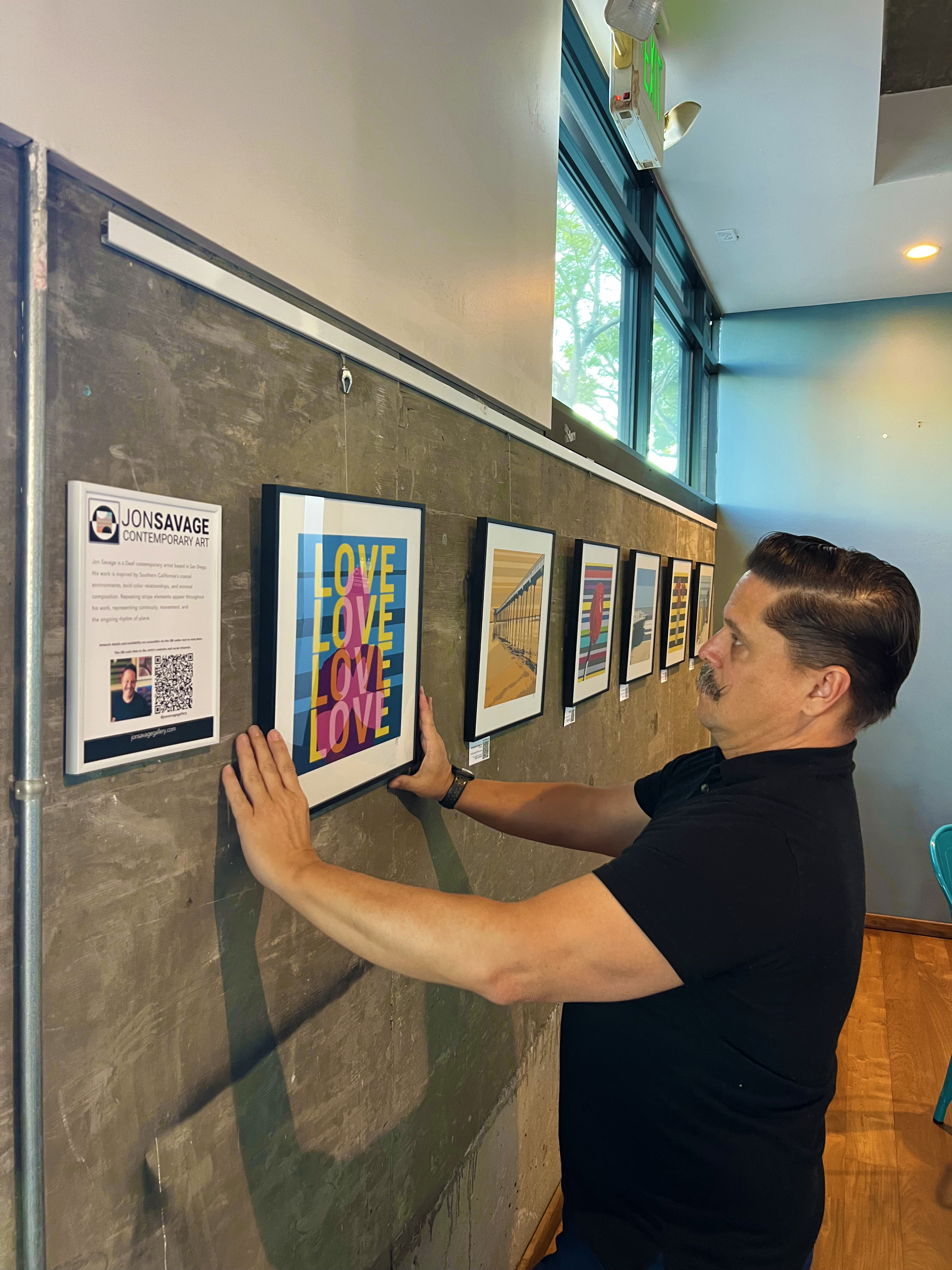 Jon Savage hand-hanging the 'Love 4 More' fine art print on a modern concrete gallery wall at Bird Rock Coffee, 2295 Kettner Blvd.
