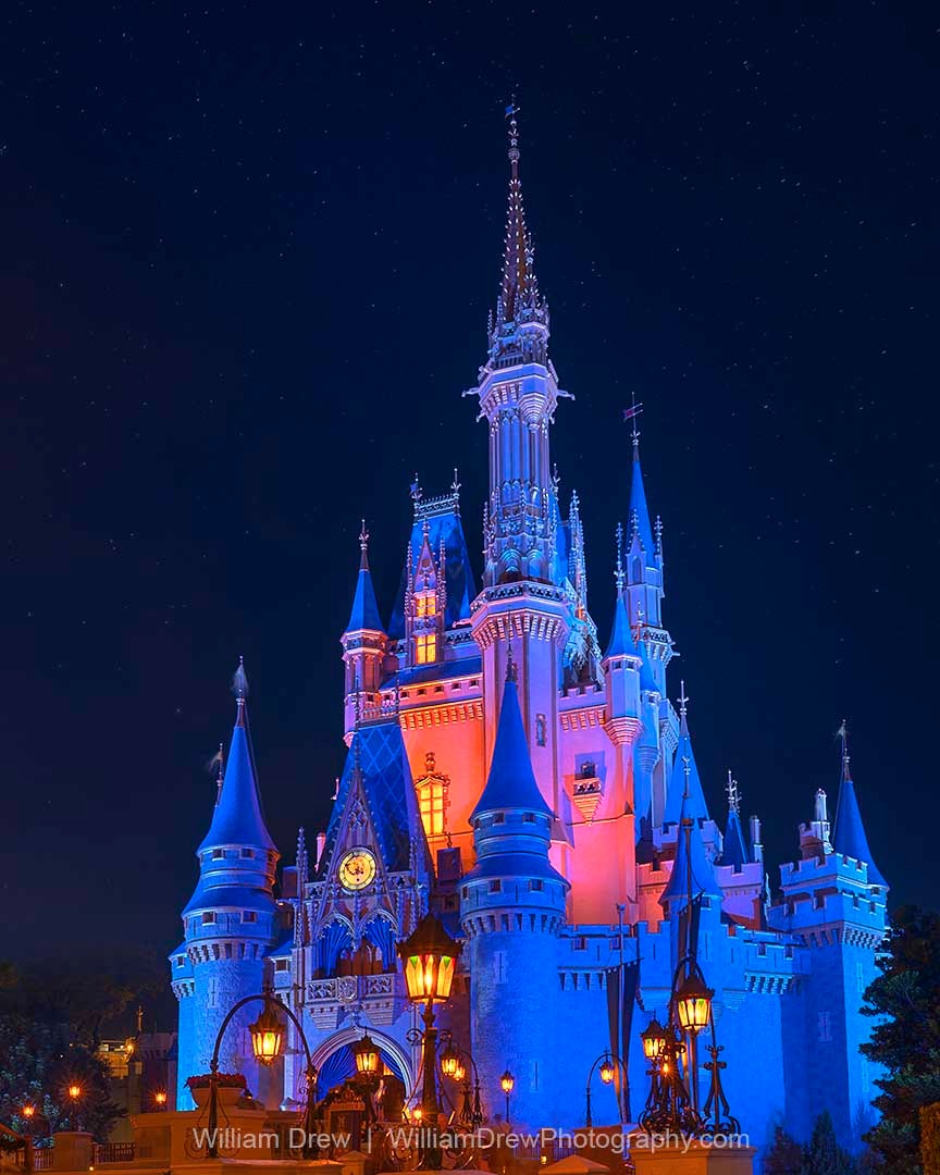 Night photo of Cinderella Castle glowing in pink and blue lights with warm lanterns and a star-filled sky at Magic Kingdom