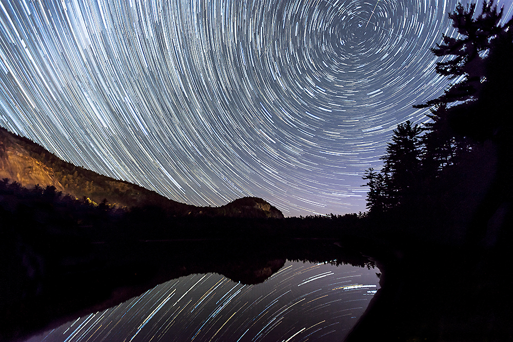 Star trails spiraling over wilderness reflected in water – cosmic memory pattern