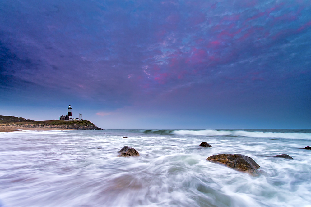 Long Island lighthouse across calm water—blue hour cools the palette while residual warm windows add contrast.