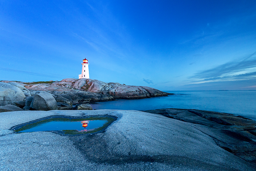 Twilight reflection of Peggy’s Cove lighthouse in a tide pool with deep blue sky and smooth granite foreground