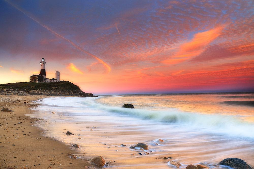 Montauk Lighthouse at sunset with waves rolling across the shoreline under vibrant orange and pink sky