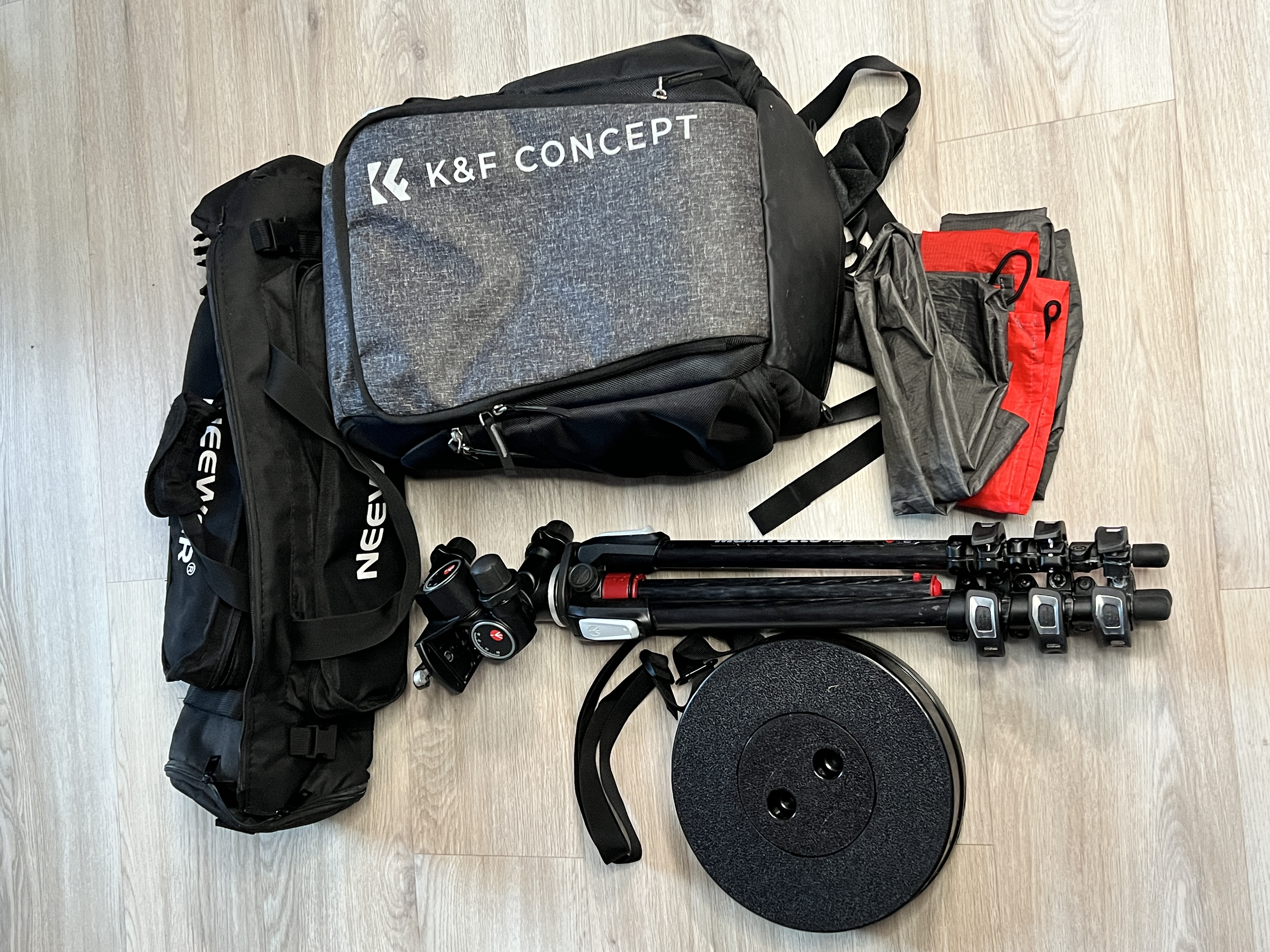A flat lay of photography gear on a wooden floor, including a black and grey K&F Concept backpack, a black bag, a tripod with a ball head, a black telescopic stool, and a folded waterproof picnic mat.