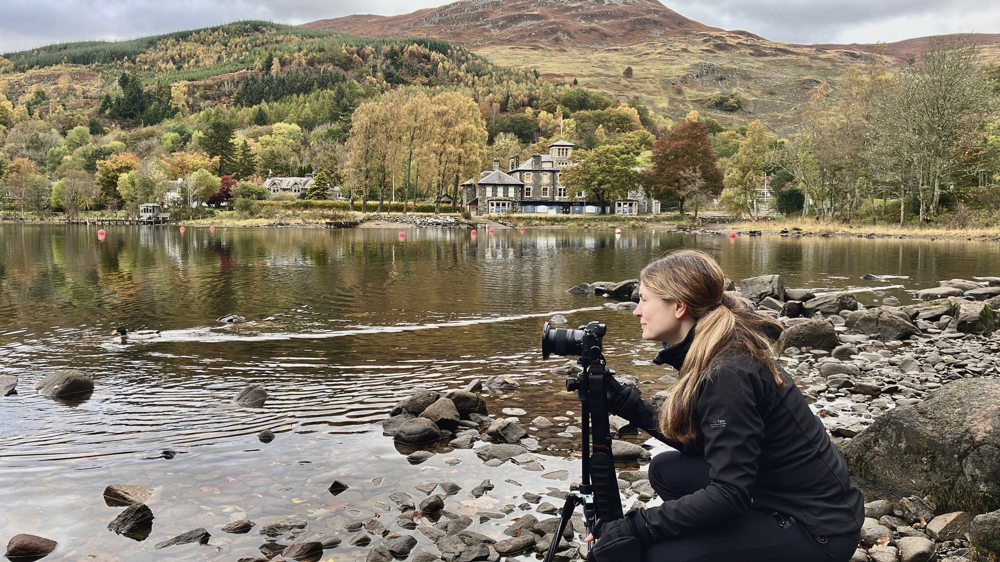 Val Miranda shown at the shores of Loch Earn with her camera shooting towards the water. On the background, some houses, autumn-coloured trees and mountains are shown.